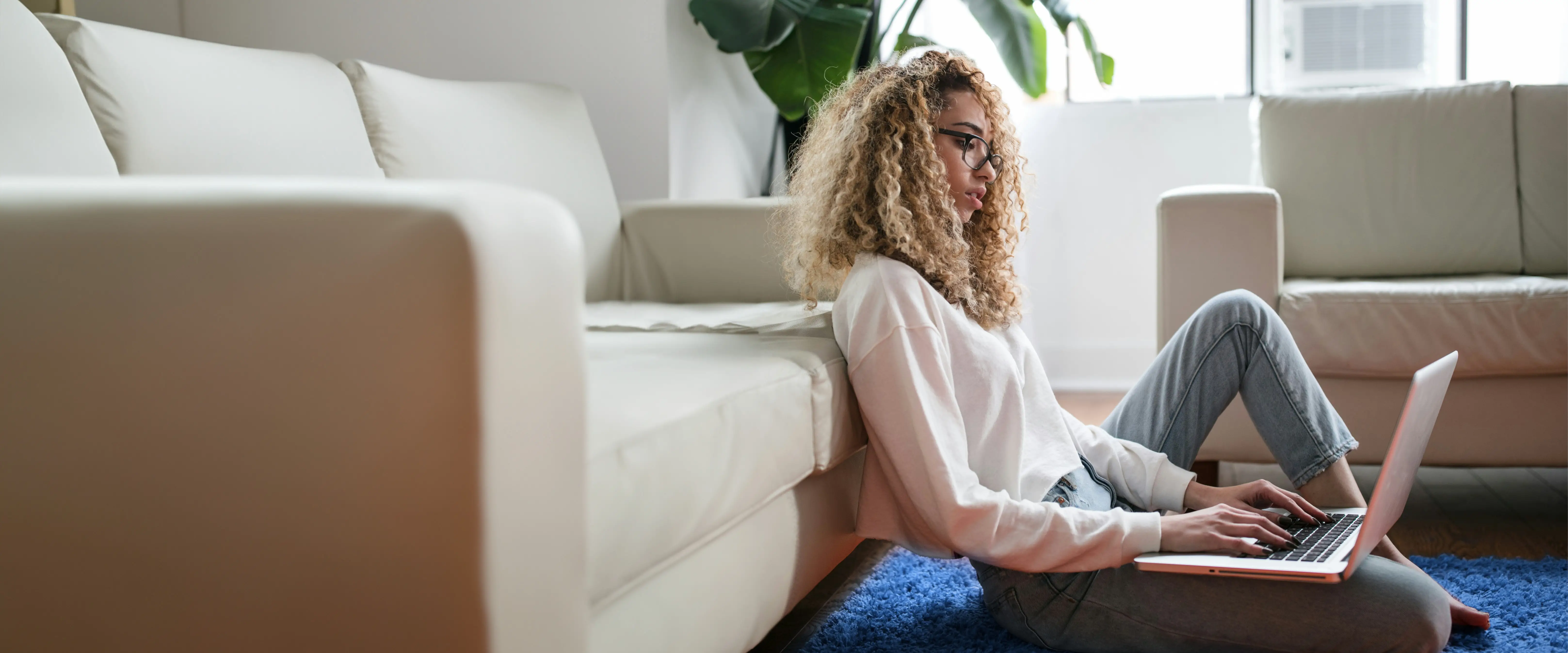 Junge Frau mit blonden Locken sitzt auf dem Teppich vor einem Sofa und arbeitet an einem Laptop.